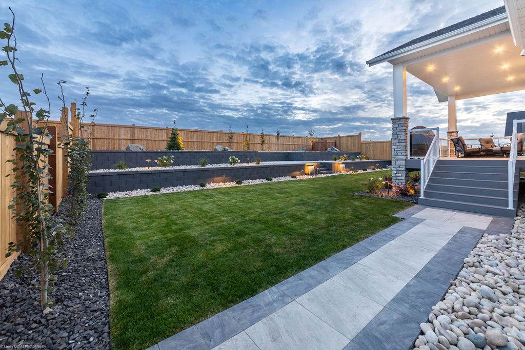 Completed backyard at dusk with a block retaining wall, lit planting beds, lawn and a paver path